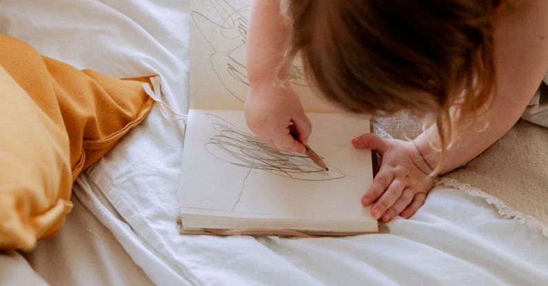 Curriculum Development - From above of small girl in dress drawing with pencil in notebook while mother using laptop on bed near