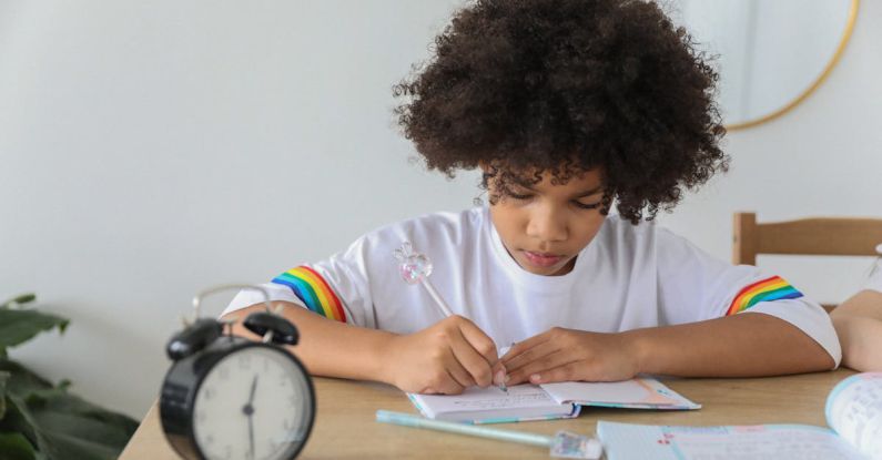 Study Time - Concentrated African American child writing in notebook while studying at desk with alarm clock at home