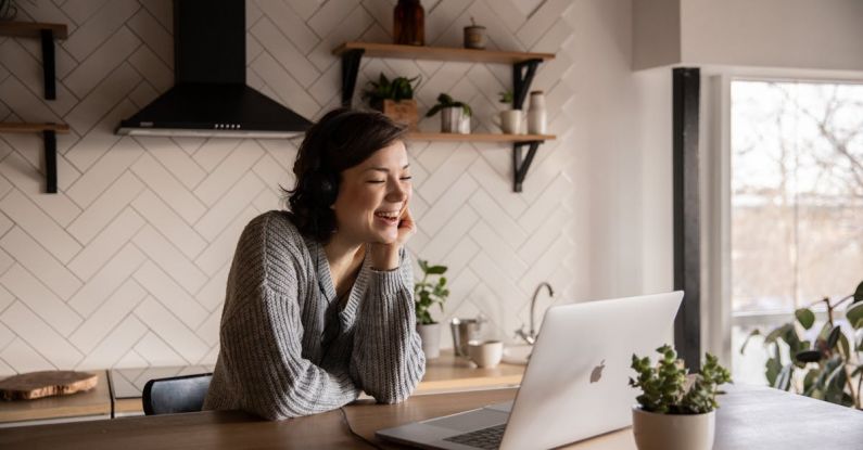 Online Discussions - Young cheerful female smiling and talking via laptop while sitting at wooden table in cozy kitchen