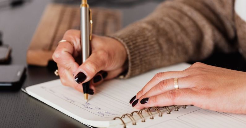 Study Plan - Crop unrecognizable female with stylish manicure sitting at black desk with keyboard and smartphone and taking notes with silver pen in notepad